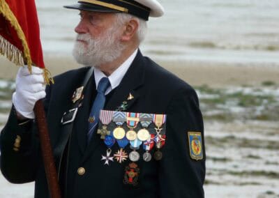 Normandy veteran in uniform with medals, holding a flag. Remembering L'Histoire du Débarquement et de la Bataille de Normandie.