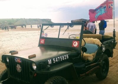 Vintage military jeep on a Normandy beach with a flag, commemorating L'Histoire du Débarquement.